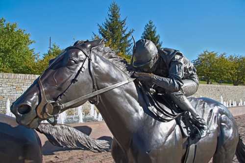 A bronze statue of a jockey riding a horse, set against a clear blue sky and green trees in the background.