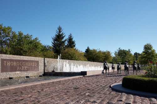 A stone wall with "Thoroughbred Park" sign, featuring horse sculptures and a fountain against a clear blue sky.