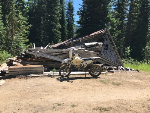 A dirt bike parked near a collapsed wooden structure in a forested area with tall trees.