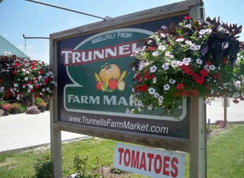 Sign for Trunnell's Farm Market featuring colorful flower baskets and a "TOMATOES" banner below.