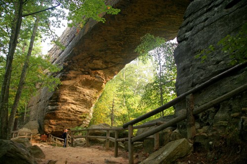 A person stands beneath a large stone arch surrounded by lush green trees and a dirt path.