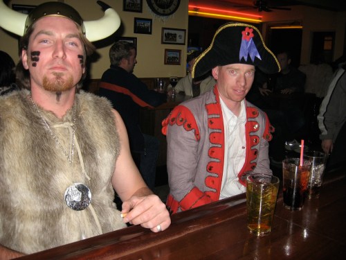 Two men in costumes at a bar: one in a Viking outfit and the other in a military-style jacket, with drinks in front of them.