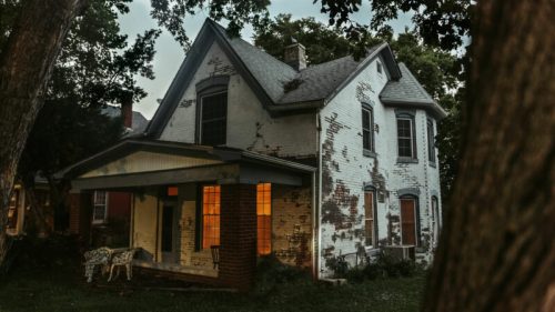 A weathered white house with peeling paint, illuminated windows, surrounded by trees in a twilight setting.