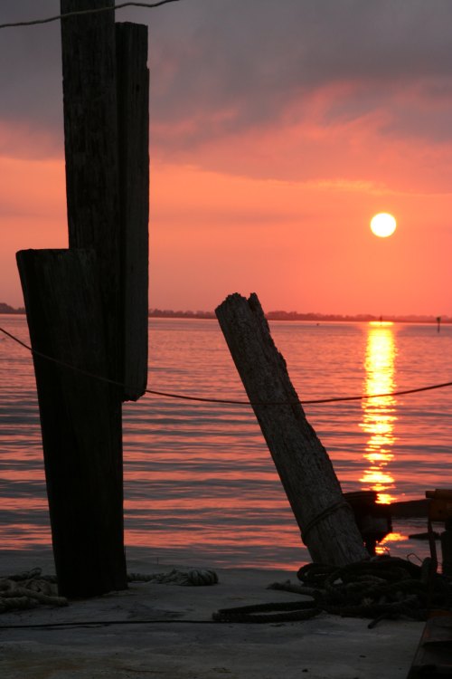 Silhouetted posts by the water at sunset, with a vibrant orange sky reflecting on the calm surface.