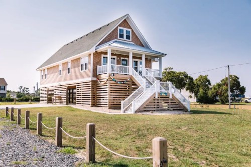 A large, two-story beach house with a wooden exterior and stairs leading to a front porch, surrounded by grass.
