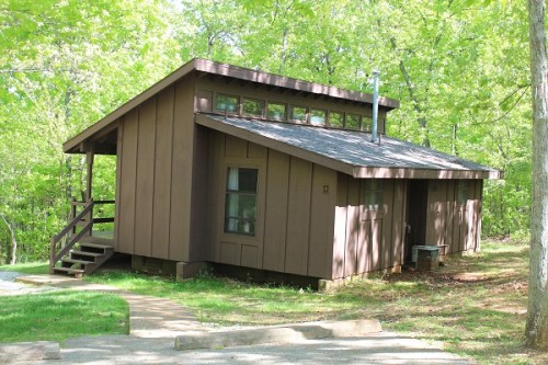 A brown wooden cabin surrounded by green trees, featuring a sloped roof and a small porch with steps.