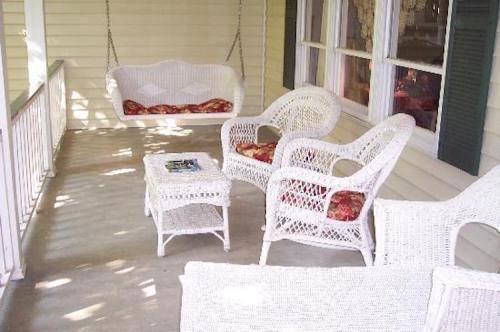 A cozy porch with white wicker furniture, including swings and a small table, surrounded by sunlight and shadows.