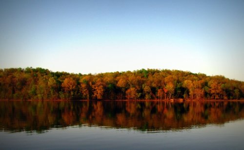 A serene lake reflecting a forest with autumn-colored trees under a clear blue sky.
