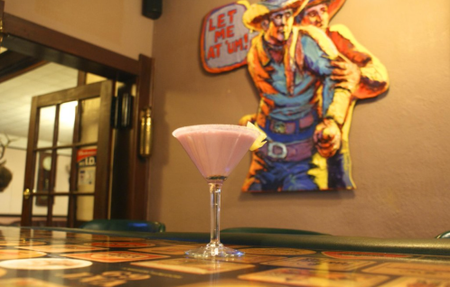 A pink cocktail in a martini glass sits on a bar, with a colorful cowboy-themed mural in the background.