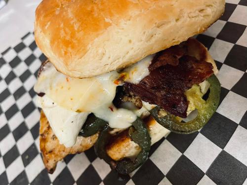 A sandwich with grilled chicken, bacon, jalapeños, and melted cheese on a toasted bun, served on a checkered paper.