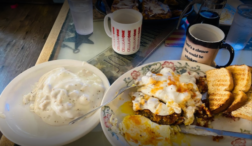 A plate of hearty breakfast with biscuits and gravy, eggs, toast, and coffee mugs on a table.