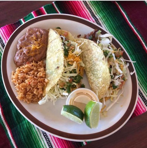 A plate of tacos with rice, refried beans, lime wedges, and a small cup of sauce, set on a colorful woven tablecloth.