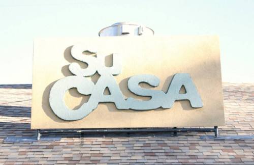 Sign on a building reading "Su Casa" against a clear sky, with a sloped roof in the background.