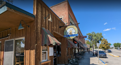 Exterior of a rustic café with a sign featuring a cartoon bee, surrounded by trees and a brick building.