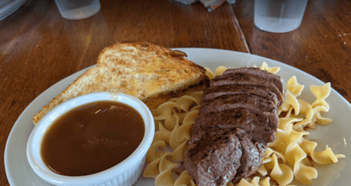 A plate with sliced beef, egg noodles, a side of gravy, and a piece of toasted bread.