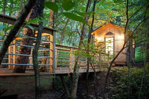A cozy wooden cabin illuminated at night, nestled among trees with a wooden walkway leading to it.