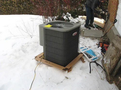 An outdoor air conditioning unit sits on a cardboard base in the snow, with a person working nearby.
