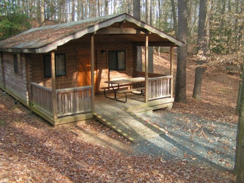 A rustic wooden cabin surrounded by trees, featuring a porch and picnic table, set in a forested area with fallen leaves.