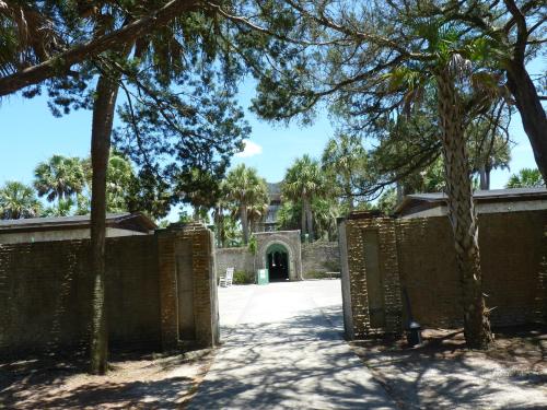 A pathway leads through stone walls to a historic building, surrounded by palm trees and greenery under a clear blue sky.