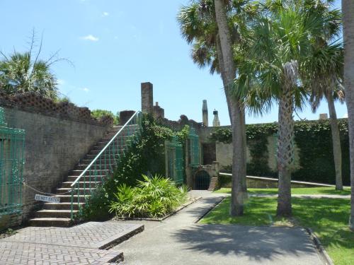 A pathway leads to brick ruins surrounded by palm trees and greenery under a clear blue sky.