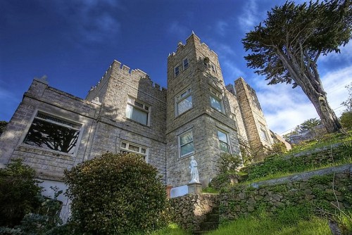 A stone castle-like building surrounded by greenery under a blue sky, featuring a statue in the foreground.