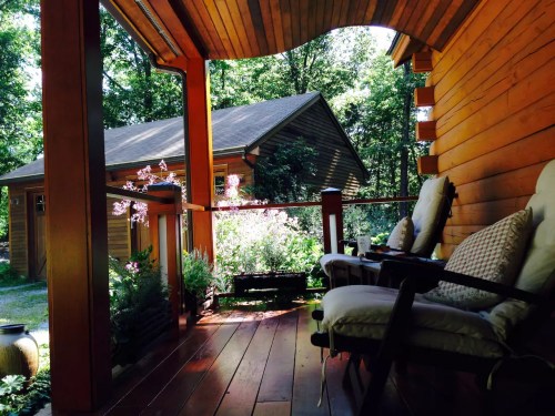 A cozy porch with two chairs, surrounded by greenery and a view of a wooden house in a serene forest setting.