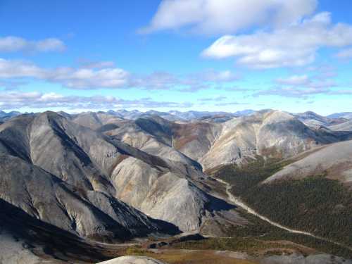 A panoramic view of rugged mountains under a blue sky with scattered clouds, showcasing varied terrain and valleys.