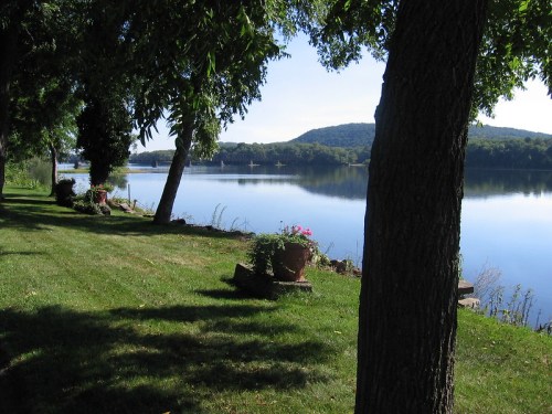 A serene lakeside view with lush green grass, trees, and a calm water surface reflecting the sky and hills.
