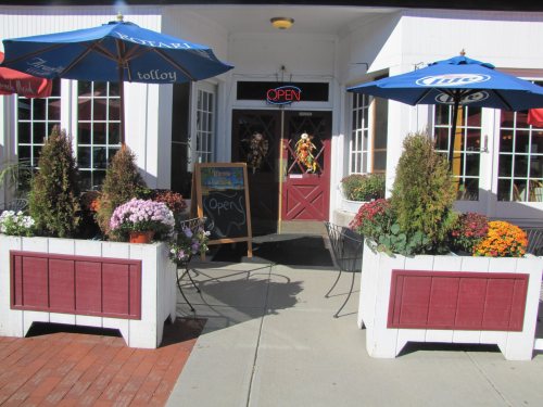 A welcoming restaurant entrance with flower planters, outdoor seating, and "Open" sign under blue umbrellas.