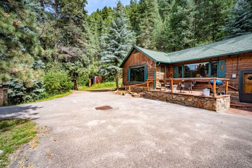 A cozy wooden cabin surrounded by tall trees, featuring a porch and a gravel driveway.