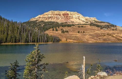A serene lake reflects a towering mountain, surrounded by evergreen trees and a clear blue sky.