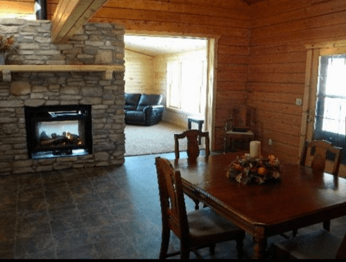 Cozy wooden interior with a stone fireplace, dining table, and a view of a living area with a black couch.