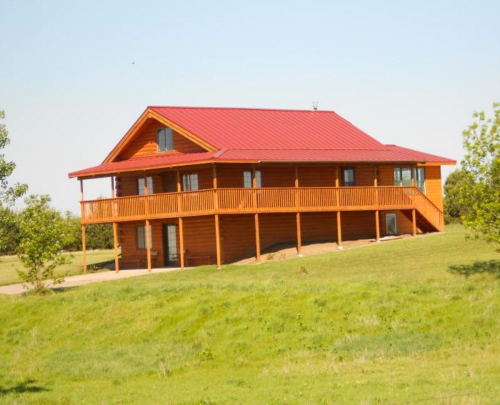 A large wooden house with a red metal roof, surrounded by green grass and trees under a clear blue sky.