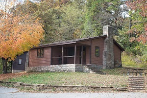 A rustic cabin with a stone chimney, surrounded by autumn foliage and a grassy area.