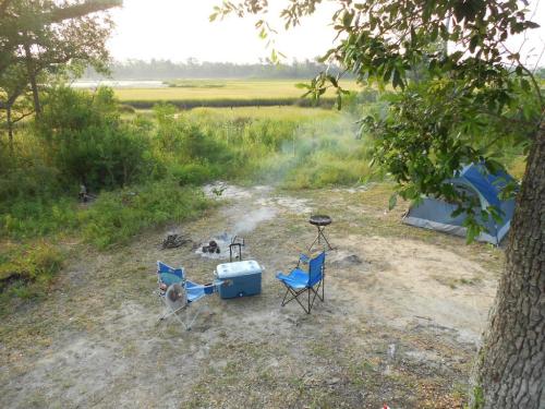 A serene campsite with a tent, chairs, a cooler, and a grill, surrounded by greenery and a view of a field.