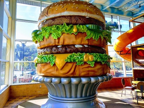 A giant, colorful hamburger sculpture with layers of lettuce, cheese, and patties, displayed indoors near a window.