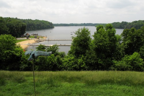 A scenic view of a lake surrounded by greenery, with a dock and boats in the distance under a cloudy sky.