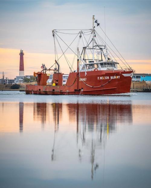A fishing boat named F. Nelson Blount anchored in calm waters, reflecting the colorful sky and nearby lighthouse.