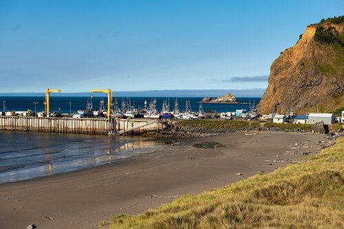 A coastal scene featuring a harbor with boats, yellow cranes, and a rocky cliff under a clear blue sky.