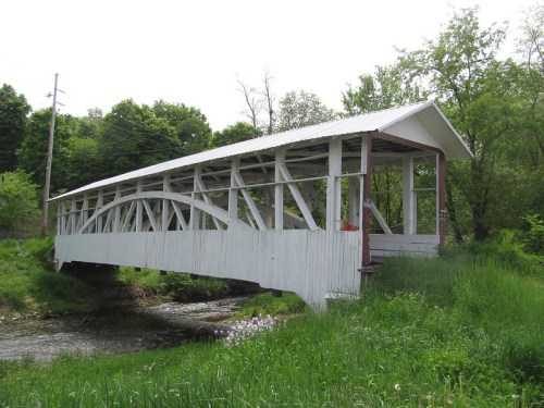 A white covered bridge spans a small stream, surrounded by lush green grass and trees.