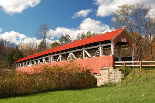 A red-roofed covered bridge surrounded by greenery and trees under a blue sky with fluffy clouds.