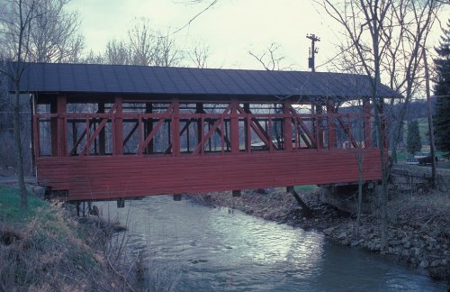A red covered bridge spans a small creek, surrounded by trees and grassy areas under a cloudy sky.