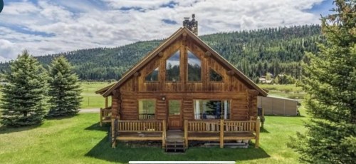 A rustic wooden cabin with large windows, surrounded by green grass and trees, set against a mountainous backdrop.