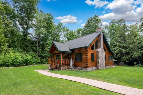 A charming log cabin surrounded by lush greenery and a clear blue sky, featuring a stone chimney and a pathway.