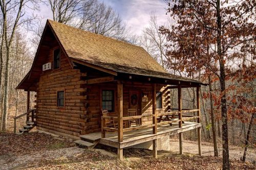 A cozy log cabin surrounded by trees, featuring a porch with chairs and a rustic, natural setting.