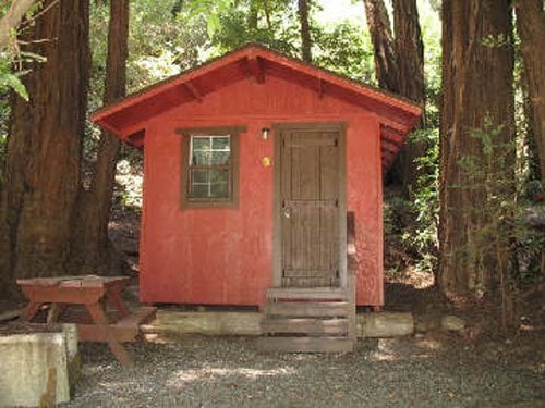 A small red cabin nestled among tall trees, with a wooden door and picnic table nearby.