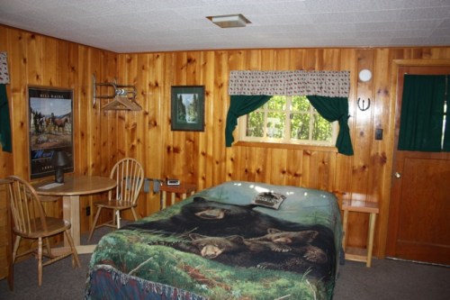 Cozy wooden cabin interior featuring a bed with a bear blanket, a small table, and a window with green curtains.