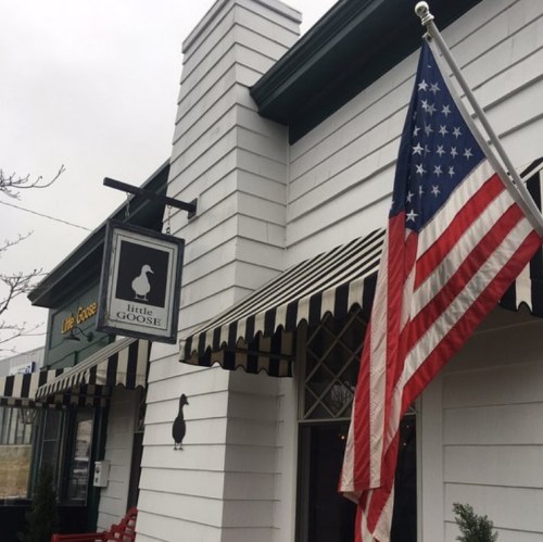 Exterior of a restaurant named "Little Goose," featuring a flag and striped awning on a cloudy day.