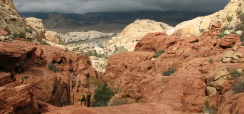 A rocky landscape with red and beige formations under a cloudy sky, showcasing a valley in the background.