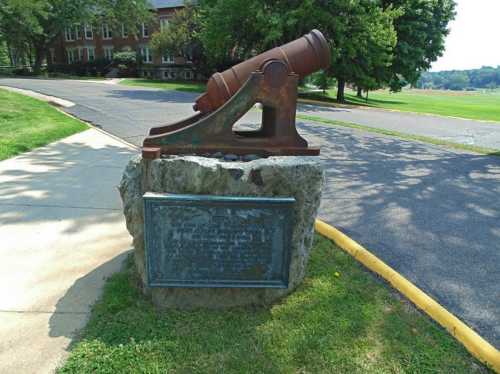 A historic cannon mounted on a stone base, with a plaque nearby, set in a grassy area near a road.